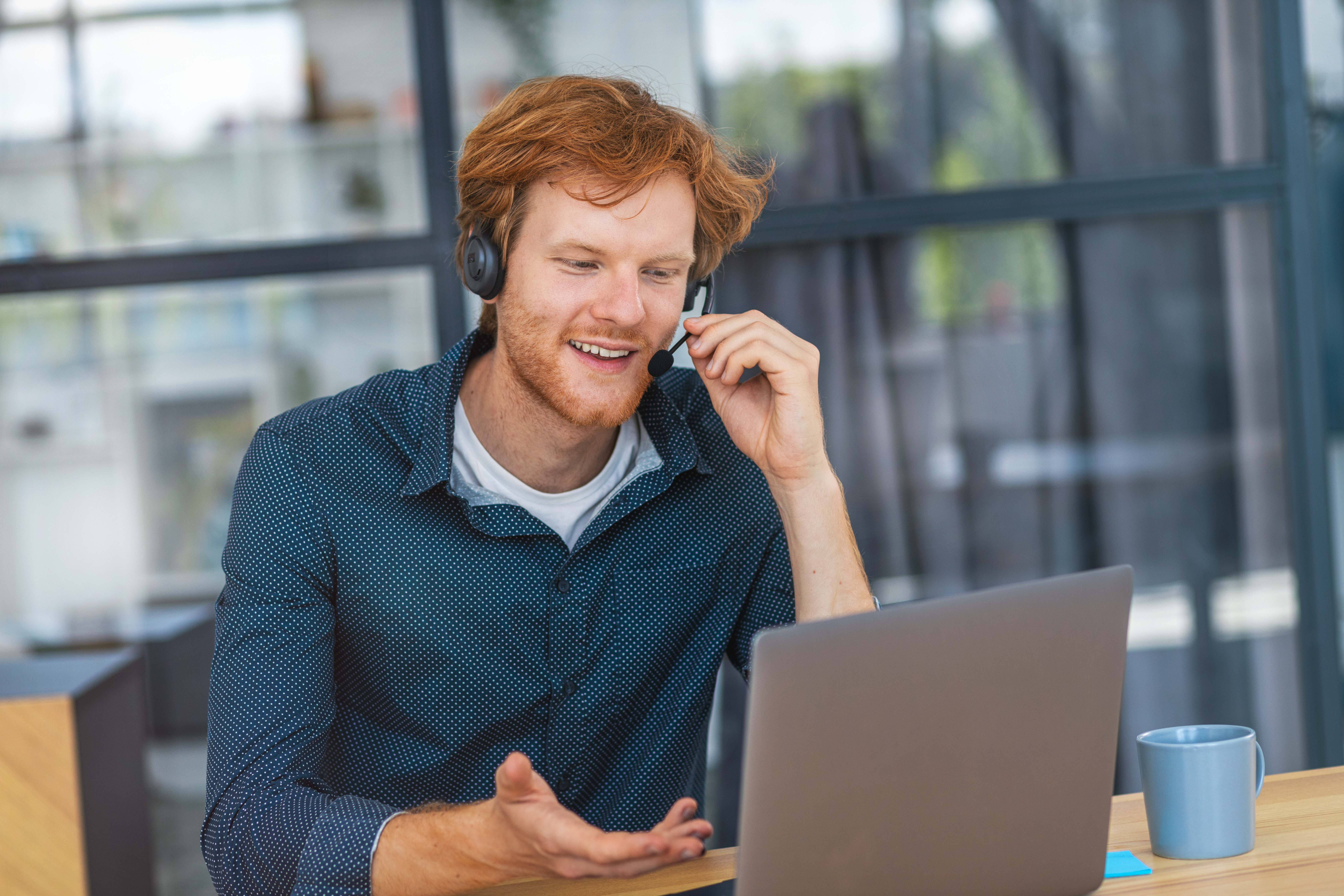 Caucasian young man customer support call center operator or receptionist sitting at the workplace in a modern office consulting a client, uses a headset, smiles friendly